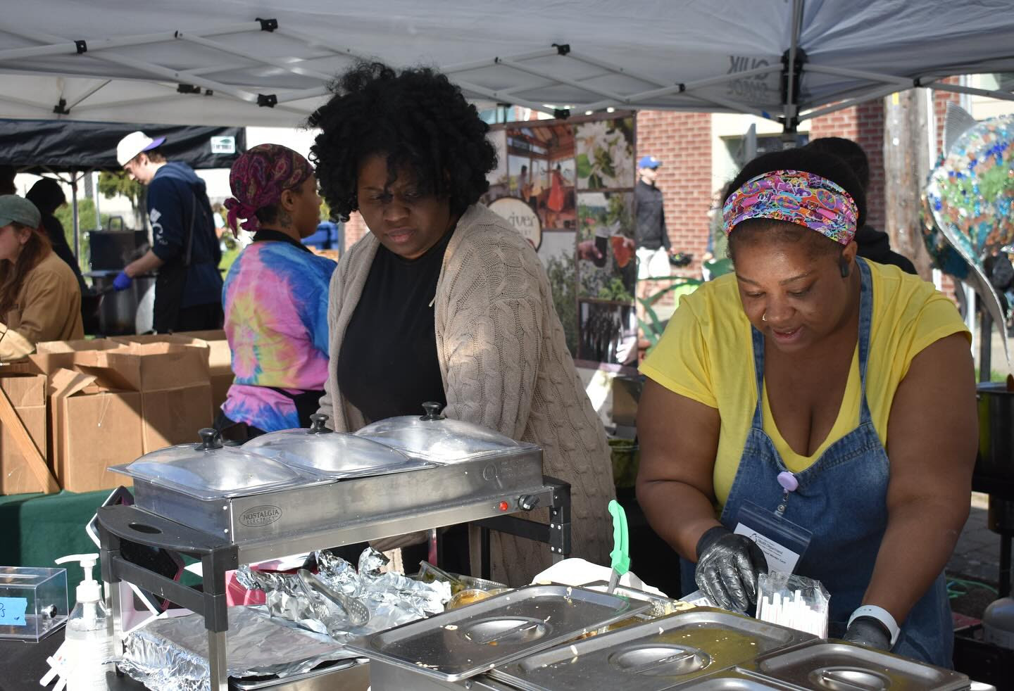 Two women prepare food under a tent at an outdoor event.